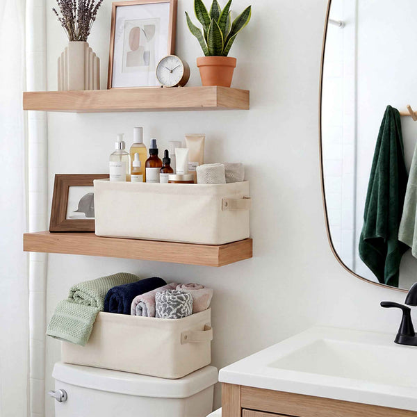Three cream canvas storage bins with wooden handles are arranged on floating wooden shelves and on top of a toilet in a bright bathroom setting.